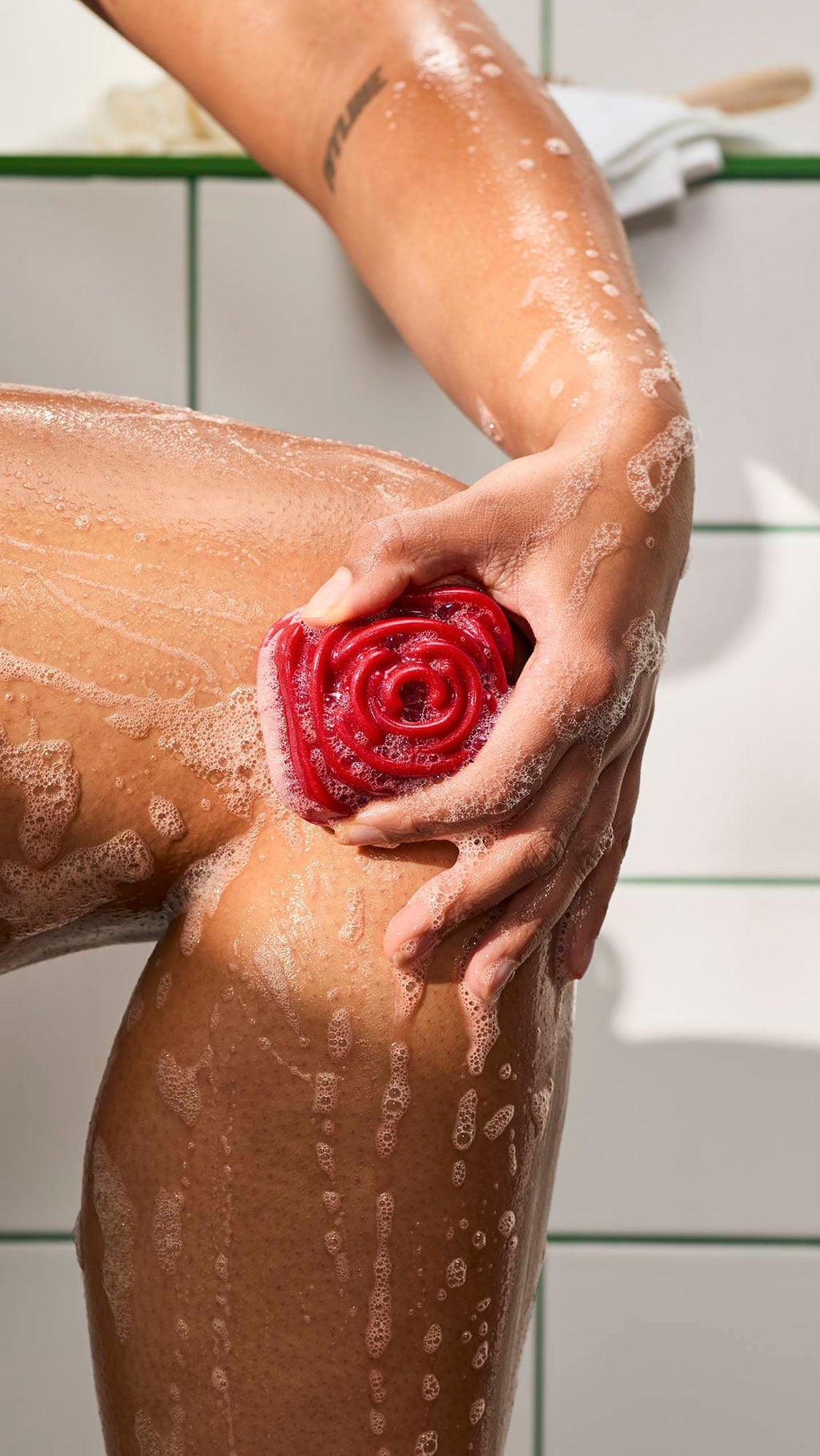 A hand lathers a wet leg with a red, rose-shaped shower gummy soap. Bubbles and foam run down the skin against white bathroom tiles with green grout.