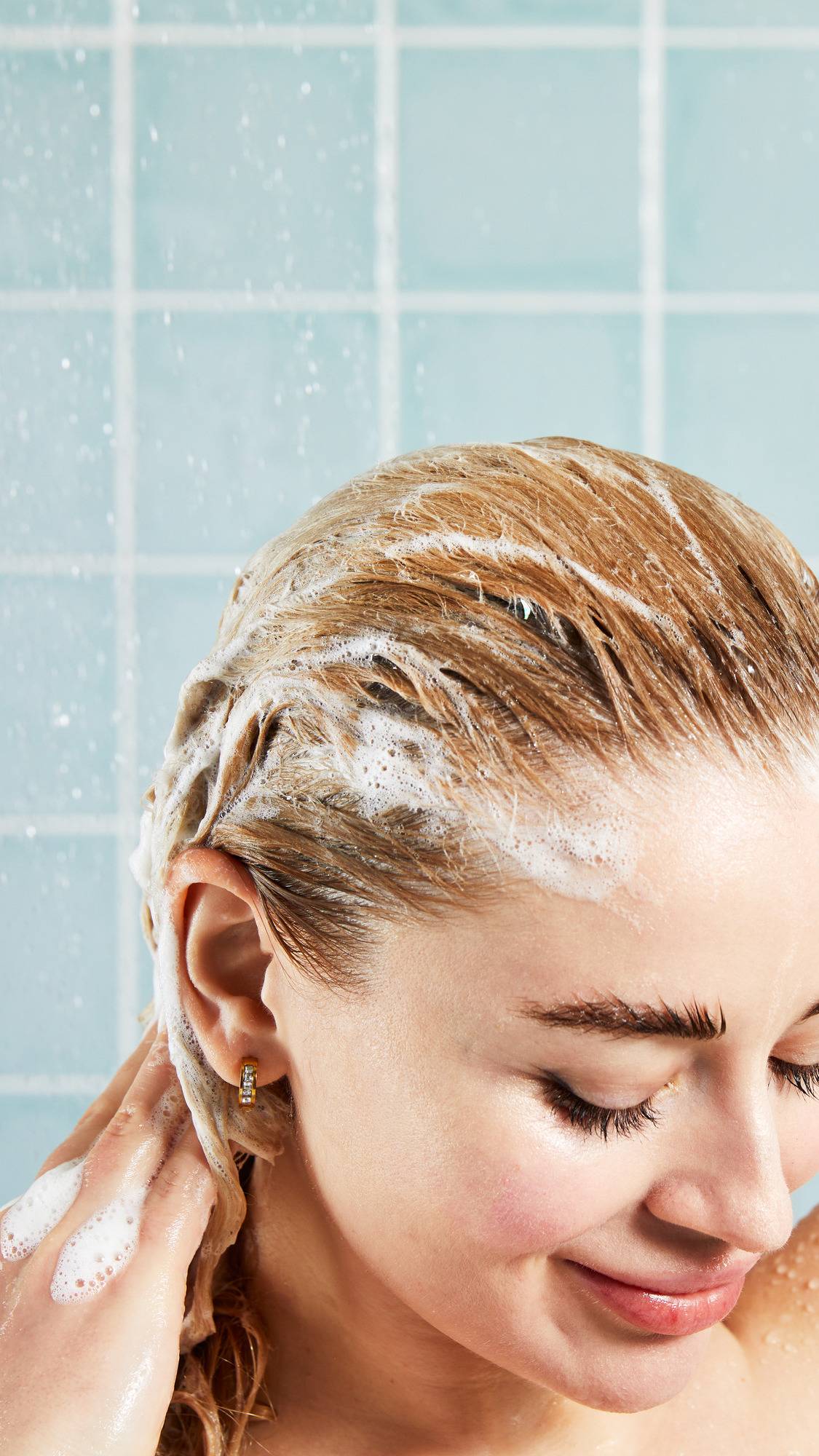 A close-up, high-angle shot of a smiling person with eyes cast down, smoothing a light shampoo lather through their blonde hair. Water droplets spray around them against a background of pale blue square tiles.