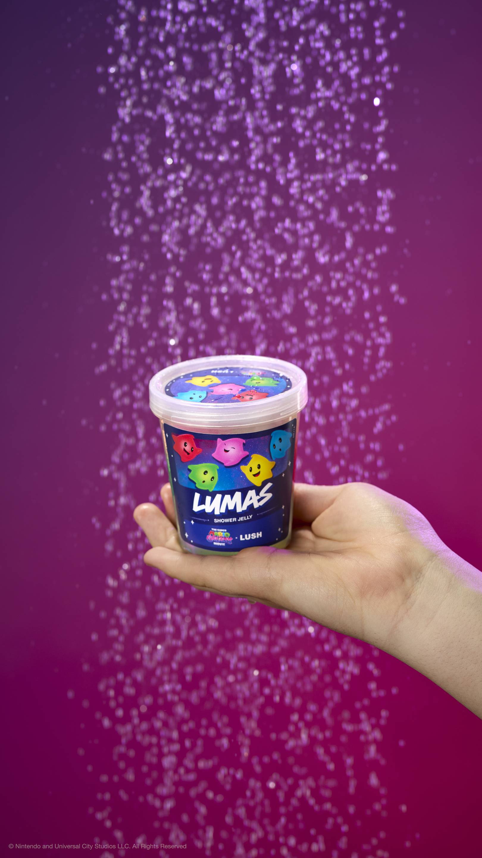 Hand holds a pot of Lumas shower jelly under a shower spray against a purple-pink gradient. Label features colorful Luma characters and movie logos. Includes plastic-free glitter.