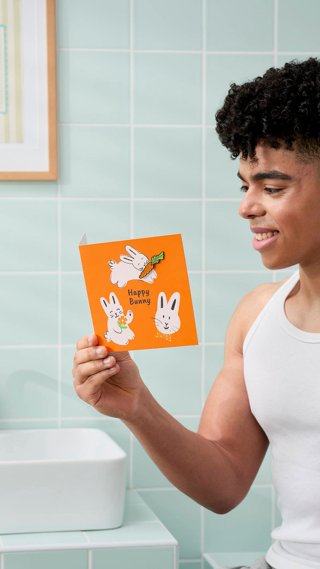 A model with dark, curly hair and a white vest smiles while holding an orange "Happy Bunny" greeting card. They are in a bathroom with pale mint-green tiles, a white basin, and a framed picture on the wall.
