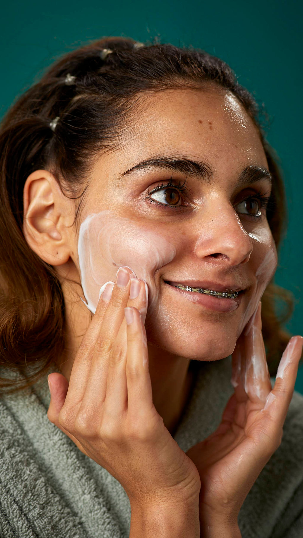A super close-up image of the model's face. They are smiling showing a glimpse of braces as they gently apply the Pineapple facial wash in circular motions over their cheek.