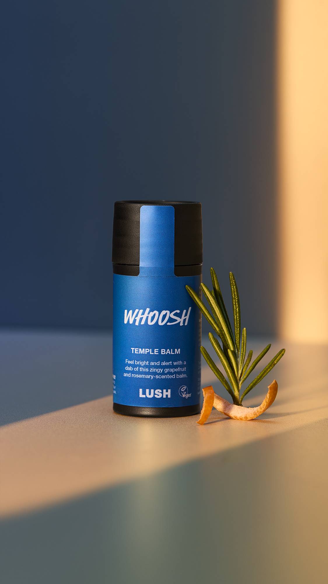 A close-up of the Whoosh temple balm surrounded by bright, natural lighting. There is a single sprig of rosemary and a twisted slice of grapefruit skin beside the product.